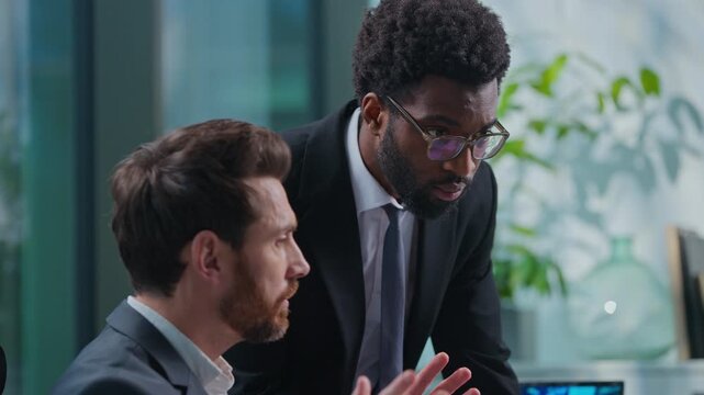 Two multiethnic business men discussing online computer job task with papers diagram businessmen talking in office African American manager asking help going away Caucasian leader work with internet