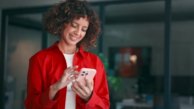 Smiling muslim arabian or indian young woman using her smartphone indoors, standing in office. Latin girl student holding mobile cell phone for texting, online banking payment or browsing. Copy space