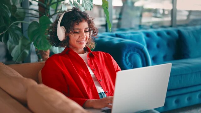 Young indian woman sitting relaxed on sofa in homeoffice using laptop computer with headphones for remote freelance work. Female curly hair teenage student girl learning studying online indoor at home