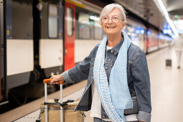 Senior woman standing on a train station platform, holding her rolling luggage and smiling, ready for a journey © luciano
