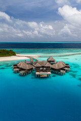Aerial view of Maldives overwater villas on stilts, thatched roofs, and infinity decks, linked by a wooden boardwalk to a palm sandbar, lagoon to cobalt ocean at midday. © Aerial Film Studio