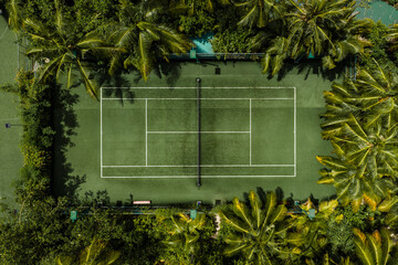 Naklejka premium Aerial view of a secluded tennis court in the Maldives, white lines and net visible, surrounded by palm trees and foliage, bright daylight and even overhead lighting.