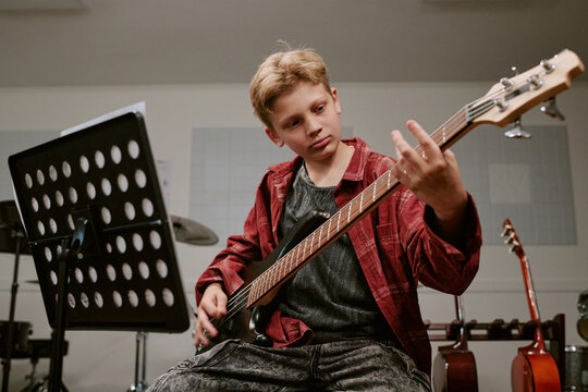 Teenager boy playing bass guitar during music lesson, practicing finger placement and performance skills. Useful for music education, youth talent, creative development campaigns