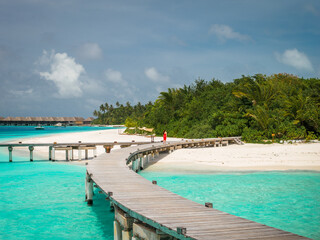Fototapeta premium A curved wooden boardwalk arcs over shallow turquoise water toward a palm island. A single person walks along it. Overwater bungalows sit on the horizon at midday.