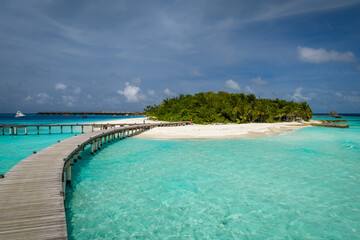 Fototapeta premium Sunlit Maldives resort shows S shaped wooden boardwalk over turquoise shallows toward white sand and palms, with overwater bungalows and a small boat under midday light.