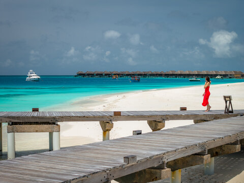 A woman walks along white sand by clear water and rustic boardwalks in the Maldives. Overwater villas extend across the lagoon and a white yacht anchors offshore at midday.