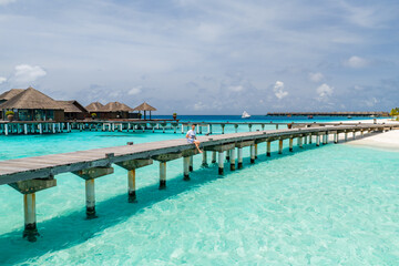 Fototapeta premium Sunlit wooden boardwalk crosses turquoise lagoon to thatched bungalows in Maldives. A person sits mid bridge, a white yacht glides on horizon, sandy beach lines right.