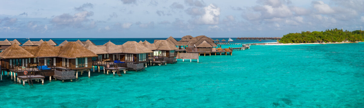 Aerial view of a Maldives island resort with thatched overwater villas on stilts, a palm covered island to the right, clear midday light, and calm Indian Ocean seas.