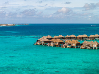 Aerial view of thatched overwater bungalows on stilts in Maldives, forming an arc by a sandy islet, calm seas and soft clouds suggest daytime tropical setting. © Aerial Film Studio