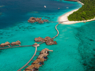 Aerial scene shows Maldives luxury resort, overwater villas, winding boardwalks, white sand beach, palm forest, thatched pavilions, boats offshore, bright daylight © Aerial Film Studio