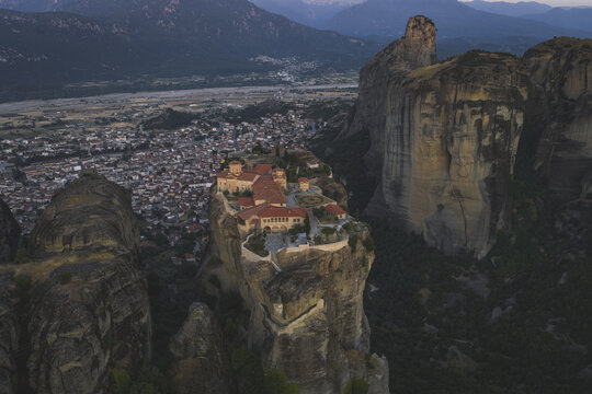 Aerial view of the historic Meteora monastery perched on a massive rock pillar with red-tiled roofs and stone walls overlooking the town and mountains in Trikala, Greece.