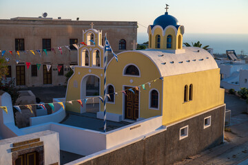 Traditional Greek Orthodox church with blue dome and bell tower at sunset on Santorini island, Greece, with festive flags and whitewashed architecture. © Andrei Tomas