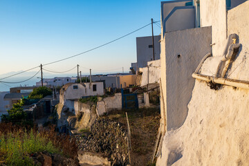 Weathered whitewashed buildings and stone walls along a quiet Santorini lane with overhead cables and distant sea views in warm evening light. © Andrei Tomas