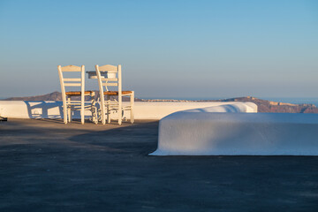 Minimalist rooftop terrace with white chairs, blue sky, and distant Aegean Sea view in Santorini, Greece. © Andrei Tomas