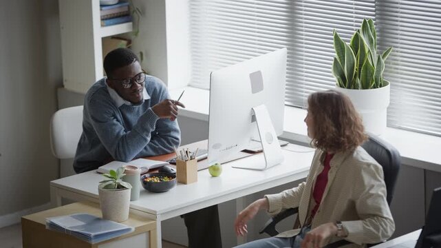 High angle shot of young adult African American man and Caucasian woman working at desks in office, discussing project. Diverse team collaboration footage ideal for corporate training videos