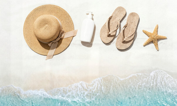 Beach essentials arranged on sand including straw hat sunscreen flip flops and starfish with ocean waves nearby.