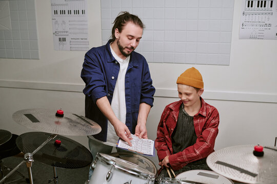 Caucasian male music teacher guiding teenage boy during drum lesson, reviewing sheet music beside drum kit. Useful for music education, private tutoring, youth learning campaigns