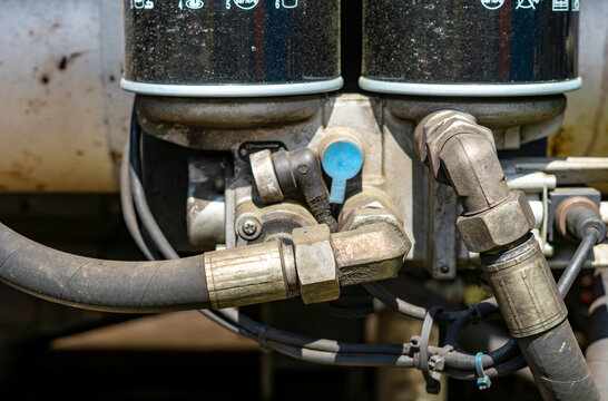 Close-up shot of a mechanical hydraulic or pneumatic system featuring two black filters on top, a metal manifold block, thick pressure hoses with metallic fittings, angled connectors, and a blue plast