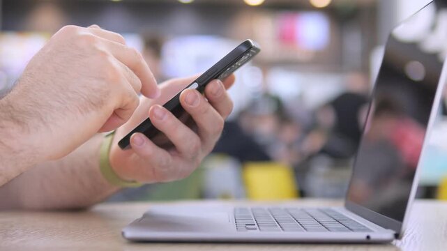 Hand holds phone, taps screen near laptop. Blurred background shows casual setting. Person focused on phone, not visible face. Ideal for tech, communication, or multitasking themes
