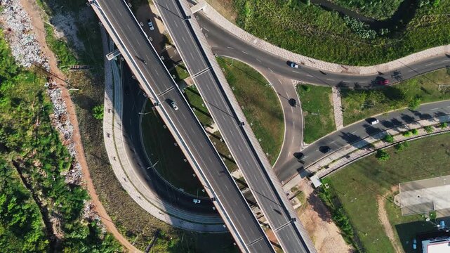Aerial view shows a complex highway interchange. Vehicles move smoothly on multiple bridges. Greenery contrasts with concrete structures. Construction sites indicate ongoing development
