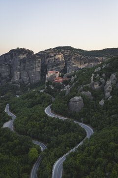 Aerial view of the Meteora monasteries perched on towering rock pillars with a winding mountain road through dense green forests at dusk in Trikala, Greece.