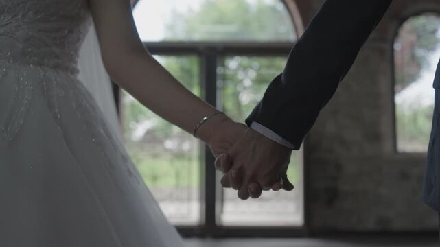 Cinematic close-up of a bride and groom holding hands. Elegant couple on their wedding day with a rustic arched window and blurred background. Love, romance, and marriage concept.