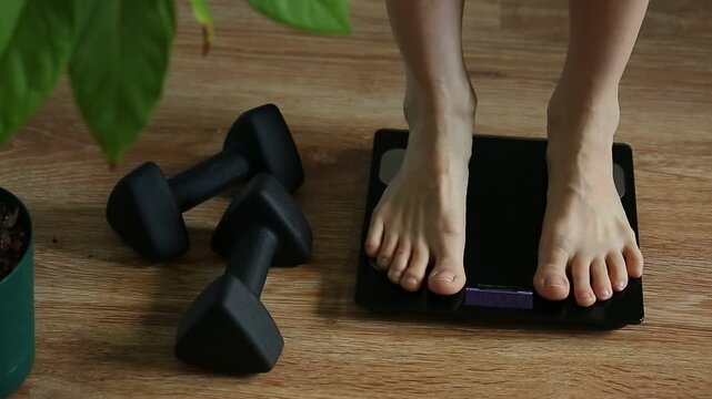 Woman bare feet stepping on black digital floor scales, she weighing, checking at home near green flower, black sport dumbbells. Measuring weight, control, wellness, fat, body care. BMI concept