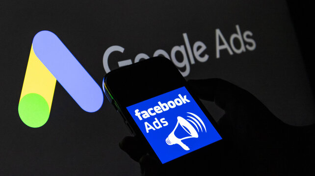 Closeup of a hand holding a smartphone displaying the facebook ads logo in front of a google ads screen