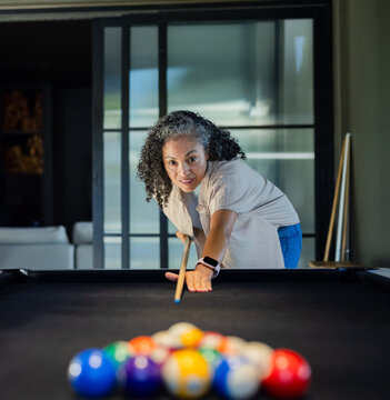 African American woman with watch leaning over black-felt table at home aiming cue at racked balls