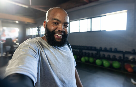 Adult African American man standing at gym wearing gray tee holding phone, taking selfie by weights