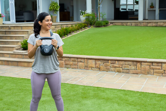 Mid-adult woman holding 5 kg kettlebell at chest while wearing workout clothes in backyard