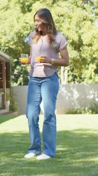 Vertical video: Walking woman from left into yard holding two orange juice glasses handing in jeans