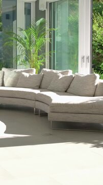 Vertical video: Panning camera revealing curved sofa at home near doors, showing sunlit floor, palm
