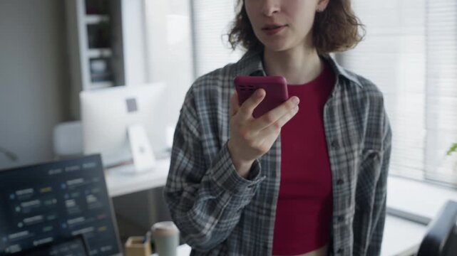 Close up of Caucasian young adult female developer using smartphone while speaking to AI assistant in tech office. Clip perfect for promoting AI voice recognition software and mobile apps