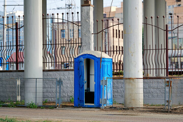 Naklejka premium Blue plastic portable toilet cabin with an open door. Public mobile restroom facility placed on city street pavement beneath massive overpass. Temporary sanitary water closet with an unlatched door