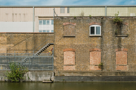 A weathered industrial building with bricked‑up windows and metal fencing stands beside a calm canal, showing the layered textures of urban history and architecture.