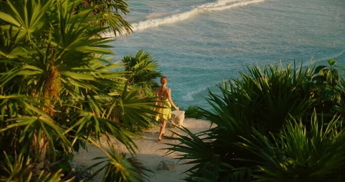 Woman in dress walking and making heart shape above tropical ocean cliff at sunset. Tropical palms frame turquoise sea in Tulum Mexico coast