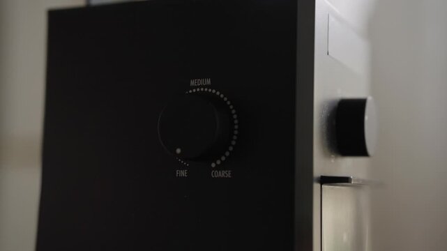 Close-up of hand turning adjustment dial on black electric coffee grinder, selecting desired grind size from fine to coarse to prepare fresh aromatic coffee beans. Shooting in slow motion