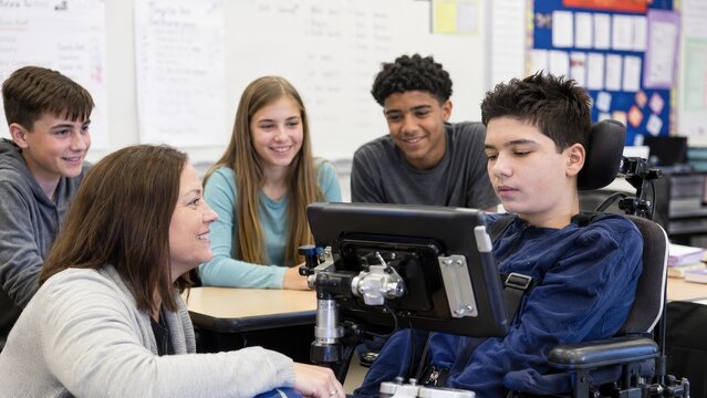 Teacher engaging with a student in a powered wheelchair using an assistive communication device while classmates watch and smile in a classroom