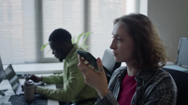 Tracking shot of young adult Caucasian woman recording voice message on smartphone in office while young adult African American man works on laptop. Footage perfect for corporate communication videos