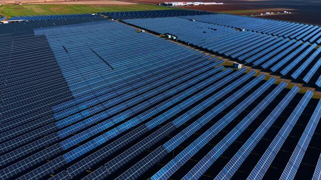 Aerial view of large solar panel farm on flat terrain. Rows of blue photovoltaic modules with inverter stations, green grass edges and brown soil under soft light.
