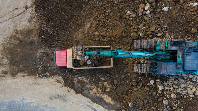 Aerial view of a teal excavator loading large rocks into a red dump truck at a construction site in Karanglewas, Central Java, Indonesia.