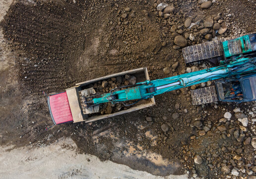 Aerial view of a turquoise excavator loading large rocks into a red dump truck at a construction site with dark soil and scattered stones Karanglewas, Central Java, Indonesia.