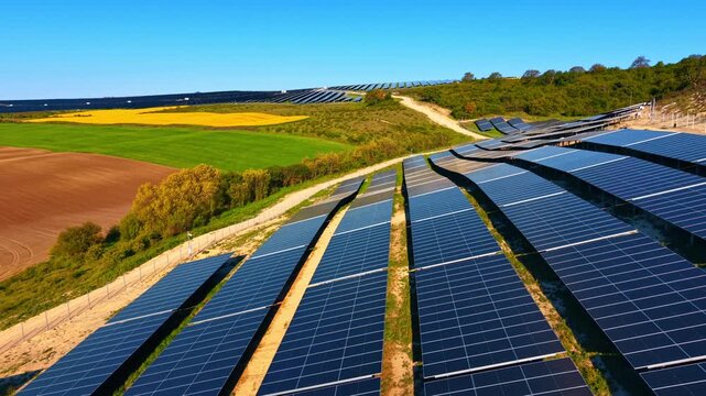 Solar panel rows on undulating terrain. Dark blue modules along a dirt path with rapeseed and green fields behind.