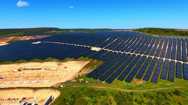 Construction site of a large solar power plant on a hillside. Excavators and equipment on cleared ground beside installed panel rows.