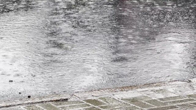 Small raindrops falling into big puddle on asphalt road. Rain drops splashing on reflective water surface. Summer rainy time. Concept of tranquility scene. Flood on heavy raining day. Rainy weather.  