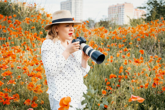 Expressive woman photographer in straw hat holding camera in poppy field near city suburbs. Urban contrast, ecology vs city expansion, creative inspiration and eco tourism concept.