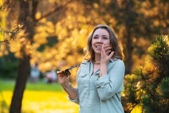 Cheerful woman laughs in park holding sunglasses. Authentic joy, vibrant summer energy.