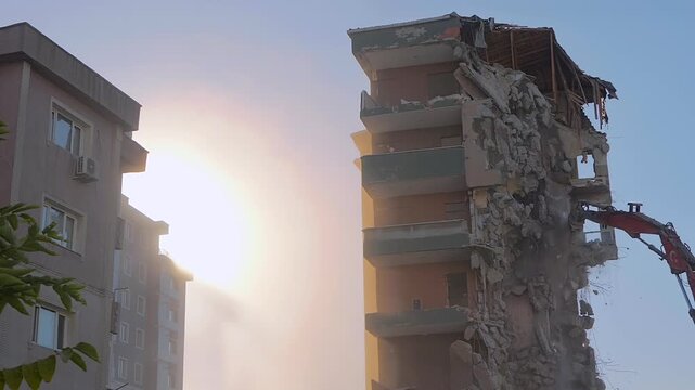 Demolition of building: excavator is tearing down the old typical Turkish multi-storey apartment, residential building in Istanbul, Turkey: slow motion. Setting sun is shining through dust