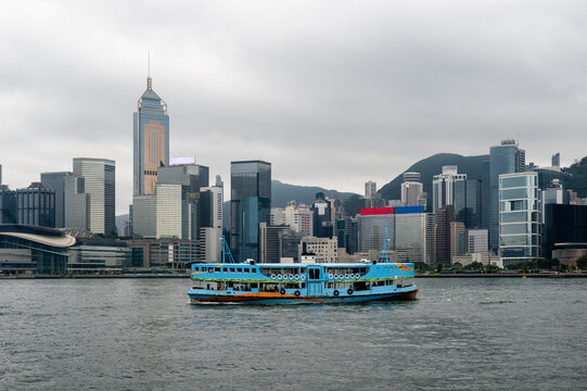 Traditional blue ferry crossing Victoria Harbour against the Hong Kong Island skyline under an overcast sky